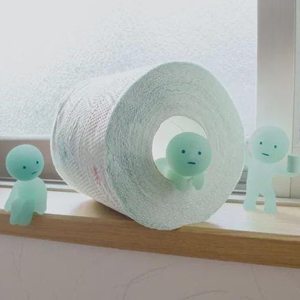 A roll of toilet paper with three small, green figurines sitting on it, placed on a windowsill.
