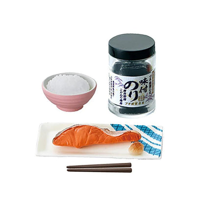 a variety of food items and utensils, including a pink bowl, a small black jar, a plate with a piece of fish, chopsticks, and a small white bowl.
