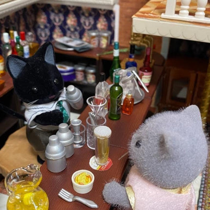 A miniature dollhouse scene depicting a bar with two toy cats, one black and one gray, sitting at a bar counter with various miniature items such as bottles, glasses, and a spoon.