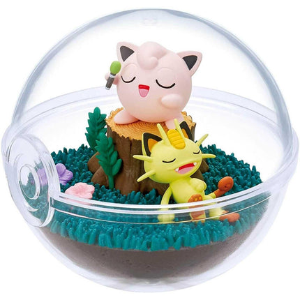 A figurine of a pink cat and a yellow cat sitting together in a glass terrarium, surrounded by green plants and a brown tree stump.