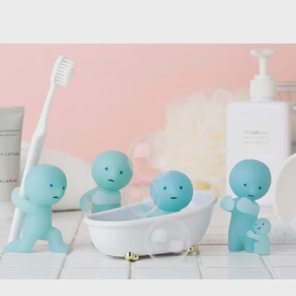 A group of four small, light blue figurines are arranged on a white tiled surface, with one figurine standing on one leg and another figurine holding a toothbrush.