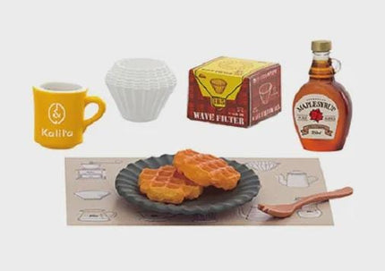 a breakfast spread, including a yellow mug, a white cup, a box of maple syrup, a plate of waffles, and a wooden spoon.