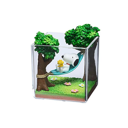A small glass container containing a miniature garden scene with a Snoopy doll lying in a hammock, surrounded by trees and flowers.