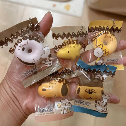 Hand holding multiple Peanuts bakery charm keychains in sealed plastic bags with silver chains