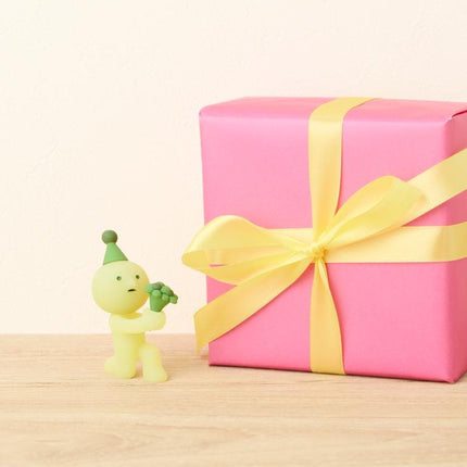 A small yellow figurine wearing a party hat is standing next to a large pink gift box with a yellow ribbon bow.