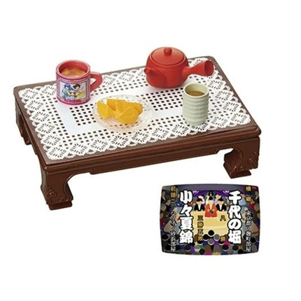 a wooden table with a lace tablecloth, upon which there are two cups, a teapot, and a plate of fruit. There is also a small box with Japanese text on it.