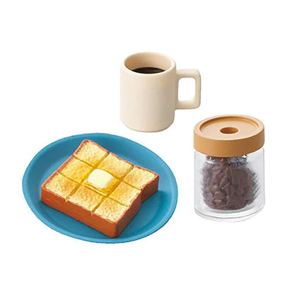A blue plate with a slice of toast, a white mug, and a glass jar filled with coffee beans are arranged on a white background.