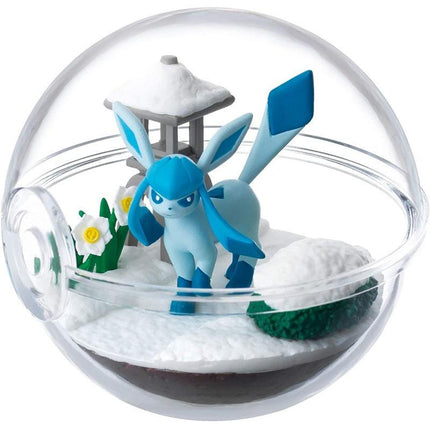 A figurine of a blue and white Pokémon character is displayed inside a clear glass terrarium, surrounded by snow, flowers, and a small structure.
