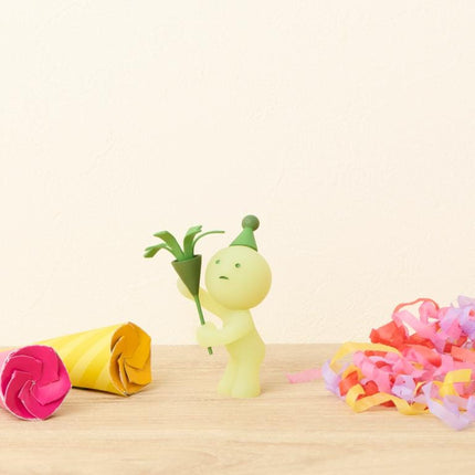 A small, green figurine wearing a party hat is holding a green flower, surrounded by colorful party streamers and a yellow paper decoration on a wooden surface.
