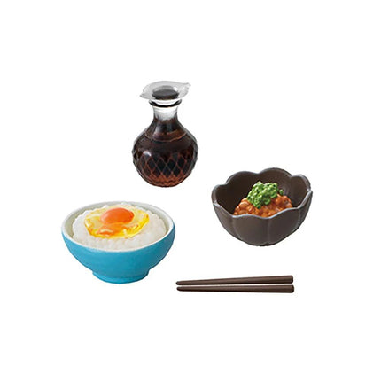 a blue bowl with a fried egg, a brown bowl with a green topping, chopsticks, and a brown bottle of sauce.