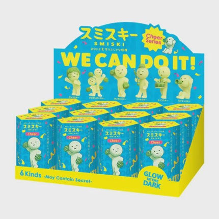 A display case containing a collection of Smiski figurines, each with a unique pose and expression. The figurines are arranged in rows, and the packaging features a bright blue and yellow design with the text "We Can Do It!" and "Glow in the Dark" written on it.