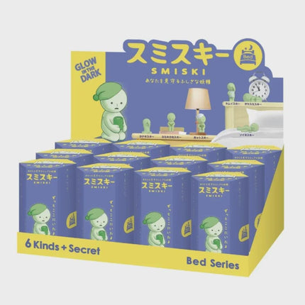 A display of glow-in-the-dark figurines of a character named Smiski, with a bed and clock visible in the background.