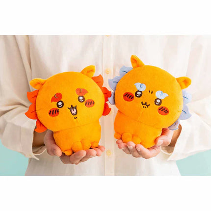 Two orange plush toys with cat-face embroidery, held in hands, colorful tufts.