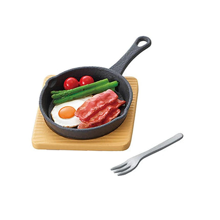 A black cast iron skillet containing a fried egg, bacon, asparagus, and tomatoes, accompanied by a fork on a wooden board.