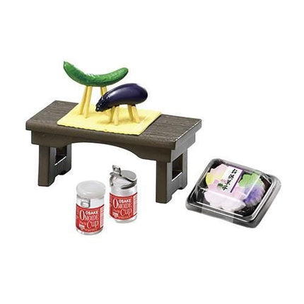 a small wooden table with various food items and containers on it. There are two small jars, one red and one clear, placed on the table. Additionally, there is a small plastic container with a lid, and a green cucumber is sitting on a yellow mat.
