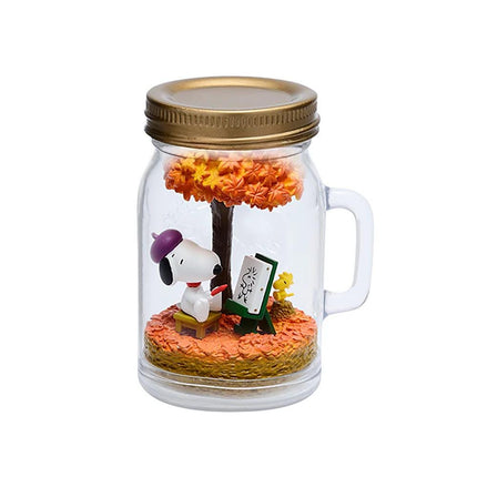 A glass jar containing a miniature scene with a Snoopy figurine sitting on a bench, drawing a picture, and surrounded by a tree and flowers.