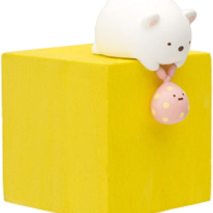 A white bear figurine is sitting on top of a yellow block, holding a pink and white polka dot balloon.
