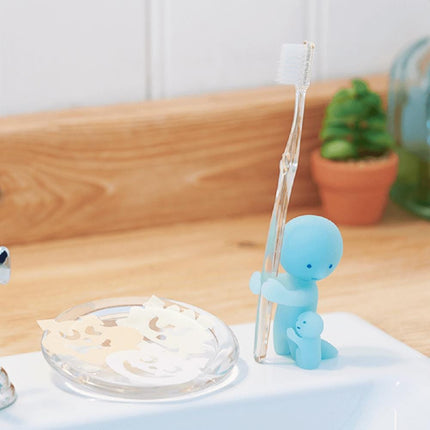 A toothbrush holder with a toothbrush and a small figurine of a child holding a toothbrush, placed on a bathroom counter.