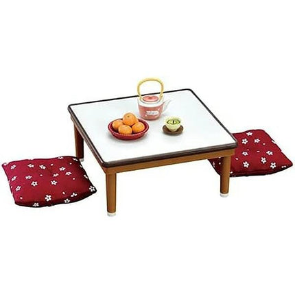 A square table with a white top, featuring a bowl of fruit, a teapot, and two red cushions with white flower patterns.