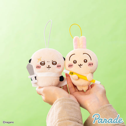 Two cream plush mascot keychains with black patches; one holds a yellow toy bar
