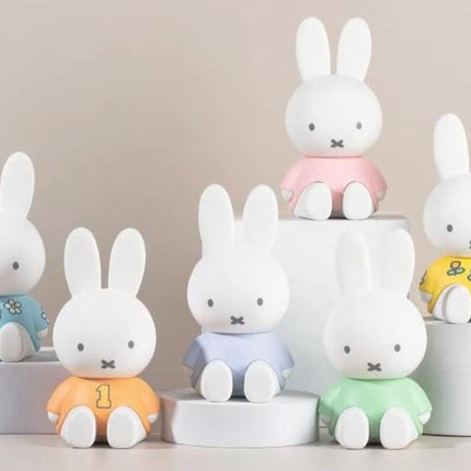 a collection of six colorful figurines of the character Miffy, each with unique outfits and poses, arranged on a white pedestal against a neutral background.