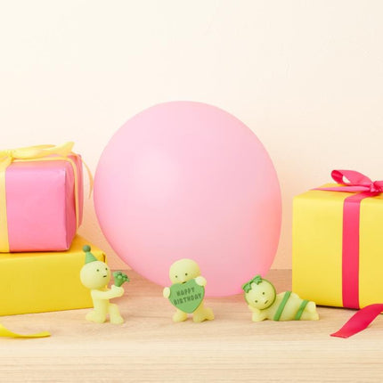 three small figurines, two of which are holding birthday cards, and a large pink balloon, all placed on a wooden surface.