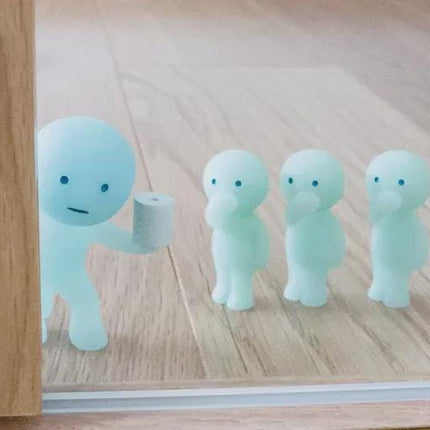 A group of three small, light blue figurines are arranged in a row on a wooden floor, with one of them holding a roll of toilet paper.