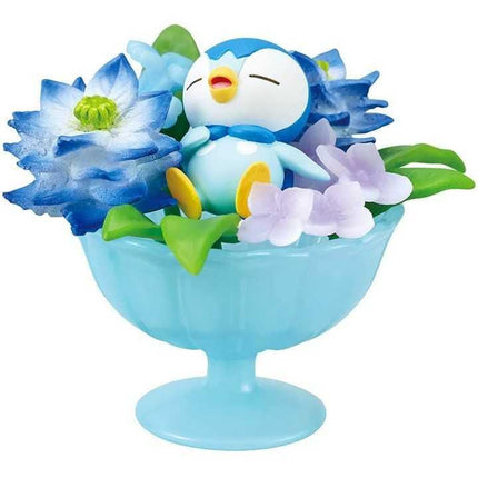 A small blue penguin figurine is sitting in a blue glass dish filled with colorful flowers, including blue and white flowers and purple flowers.