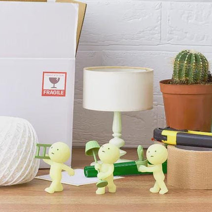 a desk with various objects, including a white cardboard box, a lamp, a cactus plant, a toy figurine, and some tape.