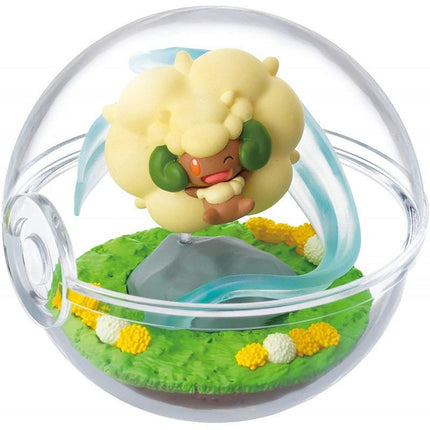 A figurine of a yellow sheep with green horns is displayed in a transparent glass ball, surrounded by a miniature landscape with grass, flowers, and a small rock.