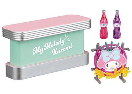 a pink and green object with the text "My Melody Kuromi" on it, along with two bottles of soda and a small toy or figurine with a pink bow.