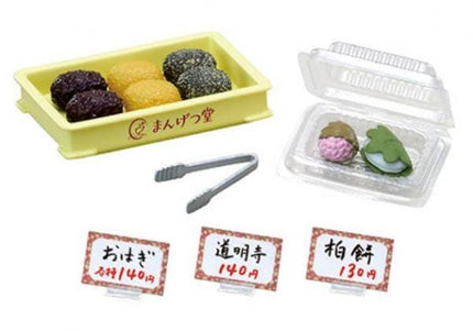 various food items and utensils, including a yellow container with food items, a clear plastic container with food items, and a pair of tongs. There are also three price tags with Japanese text, likely indicating the cost of the items.