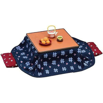 A square table with a blue tablecloth featuring a white pattern, a teapot, a bowl of fruit, and a small teacup on it.