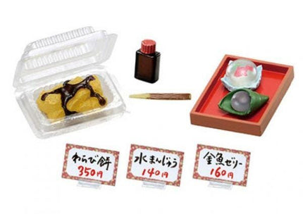 various food items and packaging, including a clear plastic container with a chocolate-covered dessert, a red tray with a white and pink food item, and three price tags with Japanese text.