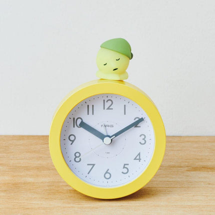 A yellow alarm clock with a small green figure on top of it, sitting on a wooden surface against a white background.