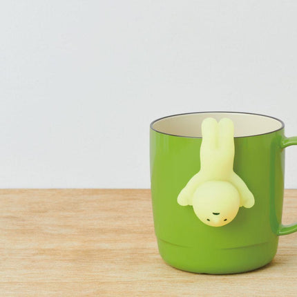 A green mug with a small yellow figurine hanging from the handle, placed on a wooden surface.