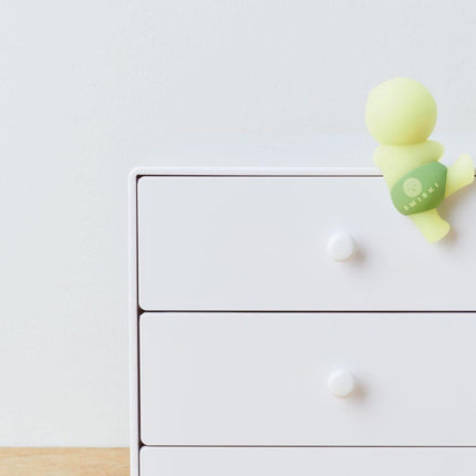 A small, yellow figurine is perched on the top drawer of a white wooden dresser.