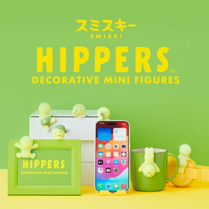 a collection of decorative mini figures, including a framed figure with a small figure on top, a green mug with a figure hanging from it, and a smartphone displaying various app icons. The background is a bright green color, and the text "Hippers Decorative Mini Figures" is prominently displayed.