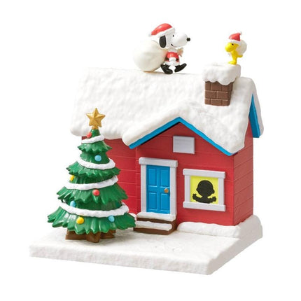 A miniature Christmas scene featuring a red house with a snow-covered roof, a decorated Christmas tree, and two small figures of characters, likely from the Peanuts comic strip.