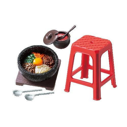 a red stool, a black bowl filled with various food items, and a small black bowl with a lid. There are also two spoons placed next to the stool.