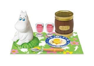 A toy hippo figurine is sitting on a green and pink floral placemat, accompanied by a wooden barrel, two glasses of red liquid, and a plate with a slice of orange.