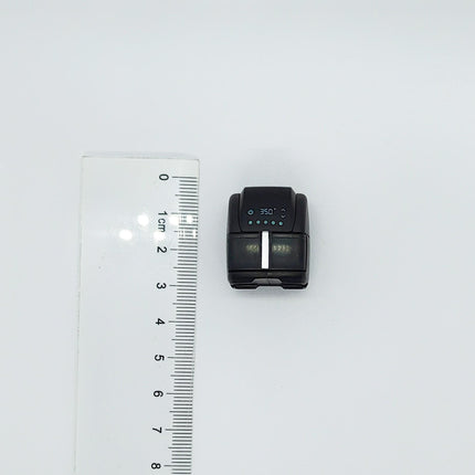 A small black electronic device, possibly a camera or a smartphone, is placed next to a ruler on a white background.