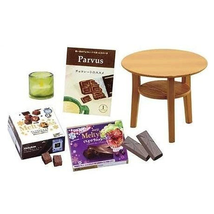 various items, including a small wooden table, a box of chocolate, a candle, and some chocolate pieces.