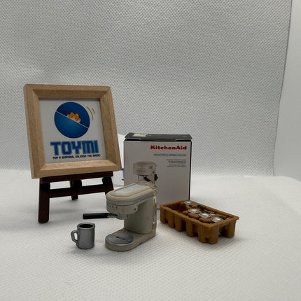 a framed sign, a box, a coffee maker, and a tray of items, all placed on a white surface.