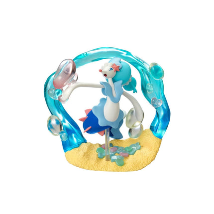 A figurine of a mermaid-like character with blue and pink colors, standing on a sandy base with bubbles surrounding it.