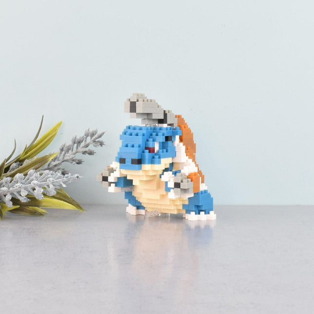 A small, colorful, pixelated figurine of a character from a video game is sitting on a reflective surface, surrounded by a few small white flowers.