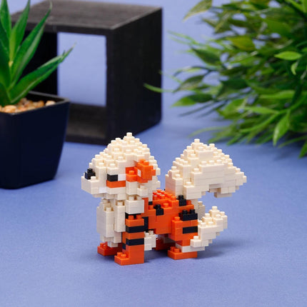 A small, colorful, and detailed toy resembling a pixelated character is displayed on a blue surface, with a potted plant and a black box in the background.
