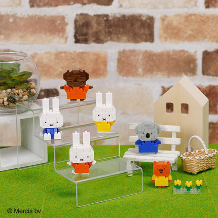 A collection of colorful, small toy animals, including a bear, rabbit, and koala, arranged on a green surface. There is also a small house and a glass container with plants in the background.
