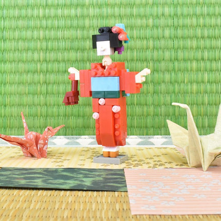 A colorful origami doll dressed in a traditional Japanese kimono stands on a woven mat, surrounded by other origami creations.