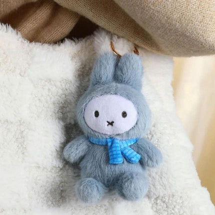 A small, plush gray rabbit toy wearing a blue scarf is sitting on a white surface.